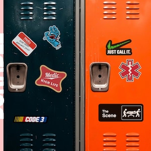 May include: Close-up of three colorful lockers with various stickers. The stickers include the text "Hello", "Medic High Life", "Just Call It", and a medical symbol. The lockers are dark teal, orange, and gray.