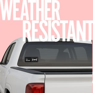 May include: A white pickup truck with a black rear window. A small black and white sticker on the window reads "The Scene" with a graphic of a person on a gurney. Large white text on a pink background reads "WEATHER RESISTANT".