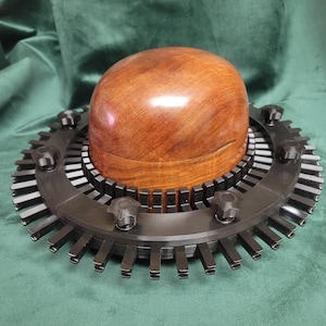 May include: A wooden hat block sits atop a black metal hat stretcher. The hat block is a light brown color and has a rounded top. The hat stretcher has a circular design with adjustable knobs and metal teeth.