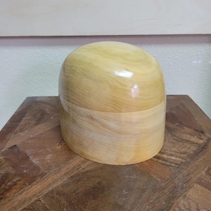 May include: A light-colored wooden hat block with a rounded top and a cylindrical base. The hat block is made of smooth, polished wood and sits on a wooden table. The hat block is used for shaping hats.