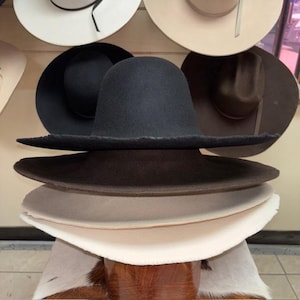 May include: A stack of various cowboy hats in shades of black, brown, and cream. The hats have wide brims and are made of felt or a similar material. Several other hats are displayed on the wall in the background.