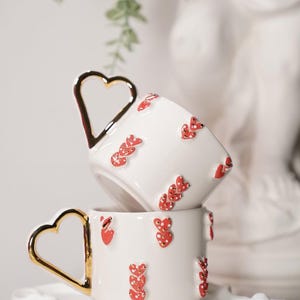 May include: Two white ceramic cups stacked on a white saucer. Each cup has a gold heart-shaped handle and is decorated with red heart-shaped designs. The cups are part of a set, perfect for serving coffee or tea.