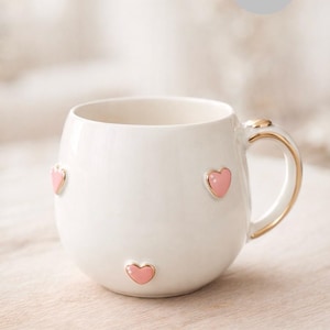May include: White ceramic mug with three pink heart accents and a gold-trimmed handle. The mug has a rounded shape and a smooth, glossy finish, perfect for enjoying your favorite beverage.