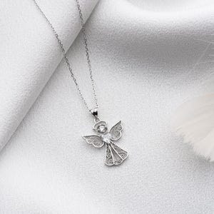 May include: A silver-colored angel pendant necklace. The pendant features an angel with detailed wings and a sparkling center stone. The necklace is displayed on a white, textured background.