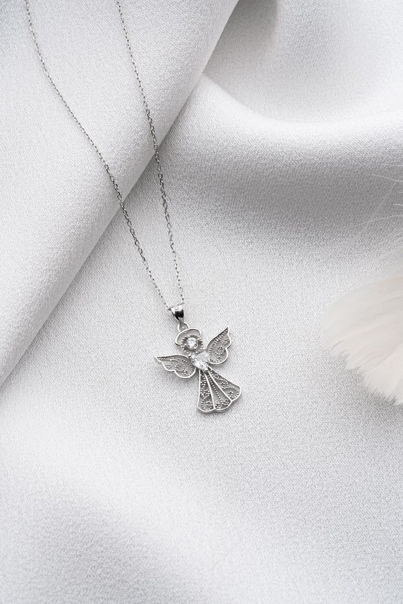 May include: A silver-colored angel pendant necklace. The pendant features an angel with detailed wings and a sparkling center stone. The necklace is displayed on a white, textured background.