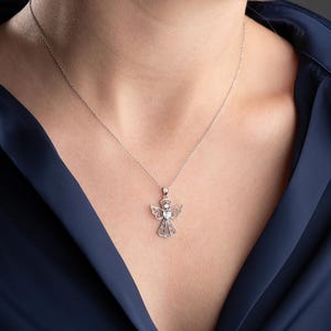 May include: A silver necklace featuring an angel pendant. The pendant has an angel figure with detailed wings and a halo, and a central gemstone. The necklace is displayed on a person wearing a dark blue top.