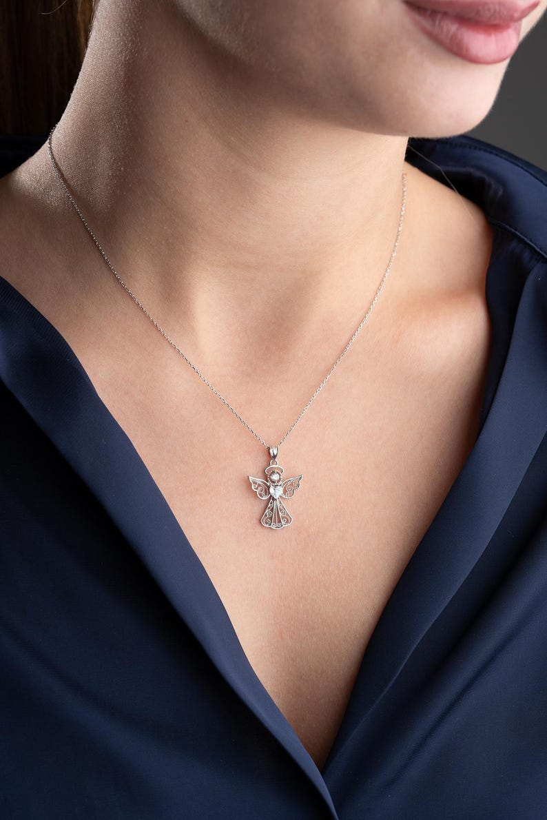 May include: A silver necklace featuring an angel pendant. The pendant has an angel figure with detailed wings and a halo, and a central gemstone. The necklace is displayed on a person wearing a dark blue top.