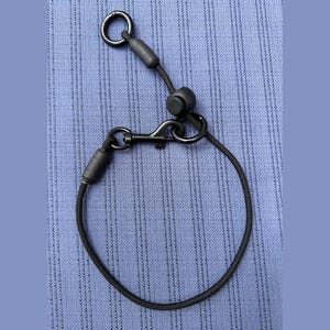 May include: A black cord accessory featuring a circular metal ring, a sliding adjustment toggle, and a metal lobster clasp. The cord is secured with two matte black cylindrical connectors, creating a circular loop design with adjustable hardware.