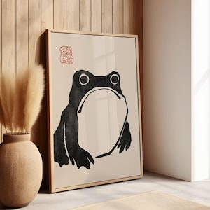 Op de afbeelding: Een ingelijste kunstprint met een minimalistische zwarte kikkerillustratie op een crèmekleurige achtergrond. De kikker heeft grote ogen en een brede mond. Een rood stempelachtig symbool staat in de linkerbovenhoek. Het kunstwerk is ingelijst.