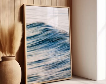 May include: A framed art print featuring a close-up of a blue and white ocean wave. The artwork is displayed in a light wood frame, leaning against a wooden wall. A beige vase with tall, dried grasses is in the foreground.