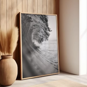 May include: A rectangular framed wall art piece featuring a black and white photograph of a large ocean wave curling and breaking. The frame is made of light-colored wood, and the image captures the texture of the water and the foam at the crest.