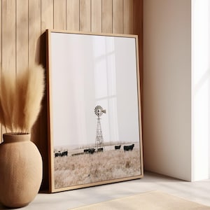 May include: A framed print of a black and white photograph of a windmill in a field with cattle. The windmill is in the center of the image and the cattle are in the background. The photograph is in a light brown frame.