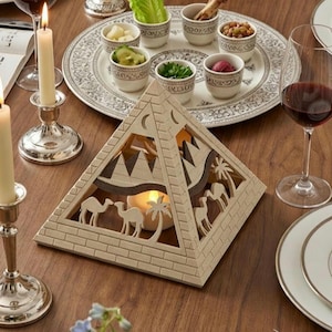 Peut inclure: Un bougeoir en bois en forme de pyramide avec des détails découpés sur le thème du désert, notamment des chameaux et des palmiers. Le bougeoir est posé sur une table en bois, avec une bougie allumée à l'intérieur. Une assiette décorative avec de petits bols de nourriture et des verres à vin sont également sur la table.