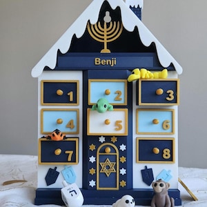 May include: A blue and white Hanukkah advent calendar shaped like a house, with eight numbered compartments and a Star of David. Includes small animal figurines and dreidels. The name "Benji" is written on the top.