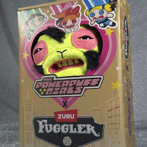 Op de afbeelding: Een boxed Powerpuff Girls Fuggler pluche speelgoed. Het pluche heeft een gele gezicht met roze accenten, zwart haar en zichtbare tanden. De doos is goudkleurig met Powerpuff Girls en ZURU Fuggler branding.