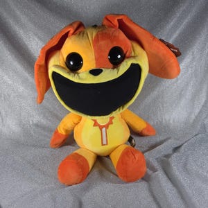 May include: A plush toy featuring a bright orange and yellow creature with large black eyes and a wide, black, toothless mouth. The character has long, floppy ears, a small black nose, and a yellow chest detail with a vertical white zipper graphic.