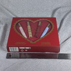 May include: A red gift set box with a heart-shaped window, containing four perfume vials. The box reads "Sweet Tooth" by Sabrina Carpenter. The vials are pink, beige, purple, and blue, each with a gold cap. A ruler is placed below the box.