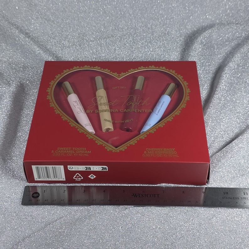 May include: A red gift set box with a heart-shaped window, containing four perfume vials. The box reads "Sweet Tooth" by Sabrina Carpenter. The vials are pink, beige, purple, and blue, each with a gold cap. A ruler is placed below the box.