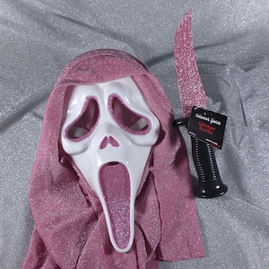 Peut inclure: Un masque Ghost Face en plastique blanc avec une capuche en tissu rose pailleté et un couteau factice rose pailleté assorti. Le couteau possède un manche noir et une étiquette indiquant Ghost Face Glitter Knife.