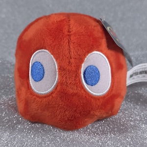 May include: A round, orange plush toy with a soft, fuzzy texture. It features two large, oval-shaped white eyes with embroidered blue pupils. A small white tag with the text BATTLE ROYALE is attached to the side of the plush, along with a dark tag.