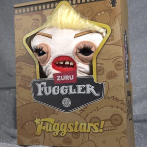 Könnte beinhalten: Ein Zuru Fuggler Plüschtier mit blondem Haar, langen schwarzen Wimpern und einem Mund mit menschenähnlichen Zähnen. Die Kreatur ist in einem braunen Karton verpackt, auf dessen Vorderseite der Text Fuggler, Zuru und Fuggstars aufgedruckt ist.