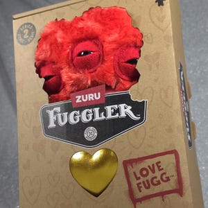 Könnte beinhalten: Ein rotes Fuggler-Plüschmonster aus der Zuru Serie 2, sichtbar durch einen Ausschnitt in einem beigen Karton. Die Verpackung zeigt ein goldenes Herz-Emblem, den Text Zuru Fuggler und den Schriftzug Love Fugg, verziert mit kleinen Herzen.