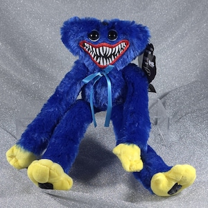 May include: A blue plush toy with long limbs, yellow hands and feet, and a wide mouth featuring sharp white teeth. The character has large black eyes and a light blue ribbon tied around its neck. A black product tag is attached to the side.