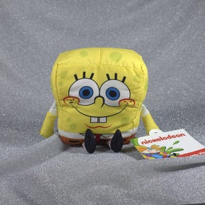 May include: A yellow SpongeBob SquarePants plush toy with a smiling face, blue eyes, and black shoes. The toy has a tag that says "Nickelodeon". The plush is sitting on a silver surface.