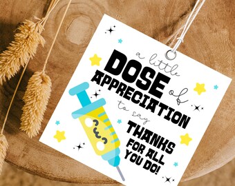 May include: A white tag with the text "a little DOSE of APPRECIATION to say THANKS FOR ALL YOU DO!" features a cartoon syringe and star accents. The tag is strung with white twine and rests on a wooden surface.