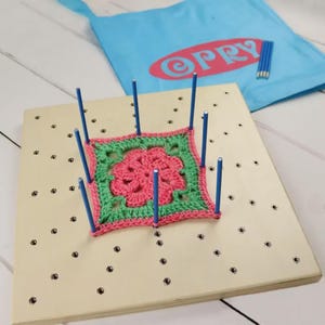 Könnte beinhalten: Ein quadratisches hölzernes Spannbrett mit einem Lochraster und mehreren eingesetzten blauen Metallstiften. Ein gehäkeltes Quadrat mit einer rosa Blumenmitte und einem grünen Rand ist über die Stifte gespannt. Ein hellblauer Beutel mit rotem PRY-Text.