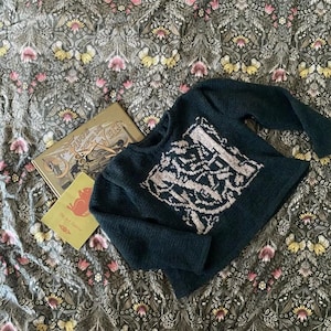 May include: A dark teal knitted jumper with a cream geometric design. The jumper is on floral fabric. Two books are next to the jumper, one titled "Hugh the Gardener". The fabric has a floral pattern.