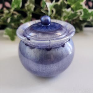 May include: A small, round, purple and white ceramic jar with a lid. The jar has a shiny, iridescent finish.