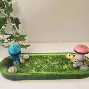 May include: A green resin tray with a mossy green surface and a blue and pink mushroom figurine. The tray is decorated with small white flowers, a bee figurine, and small stones.