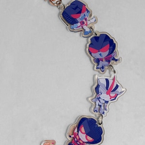 Puede incluir: Un llavero de acrílico de varias piezas con cuatro personajes robot de estilo chibi en tonos azul, morado y rojo, unidos por anillas plateadas a un cierre en forma de estrella de color oro rosa y una pequeña pieza accesoria separada.