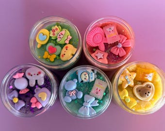 May include: Five small, clear jars filled with colorful, decorative items. Each jar contains a different color of gel and various miniature charms, including hearts, bows, and stars. The jars are arranged on a purple surface.