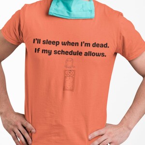 May include: A man wearing a light blue surgical cap and a light orange t-shirt with the text "I'll sleep when I'm dead. If my schedule allows." and a small illustration of a clock with a tombstone on top. He is also wearing a light blue surgical mask and light blue scrubs.