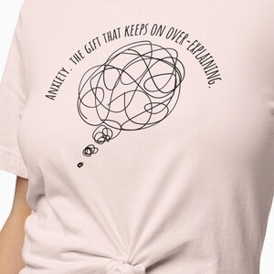 May include: A pink t-shirt with a black graphic that says "Anxiety, the gift that keeps on over-explaining." The graphic is a thought bubble with a tangled line inside.
