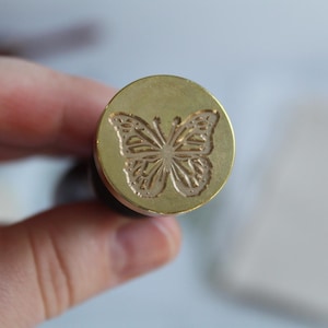 May include: A gold-colored metal stamp with a detailed butterfly design. The butterfly is centered on the stamp and has intricate wings.