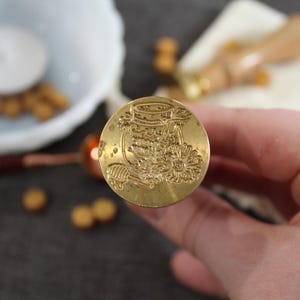May include: A gold-toned metal wax seal stamp with a detailed design of a jar with a flower and a bee. The stamp is held in a hand.