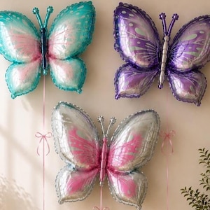 May include: Three butterfly-shaped foil balloons in teal and pink, purple and silver, and silver and pink. Each balloon features detailed wing patterns and thin ribbons hanging from the bottom, displayed as a set of decorative party inflatables.