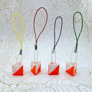 May include: Four orange and white orienteering control flags with different coloured strings attached. The flags are hanging from a white surface with a topographic map pattern.