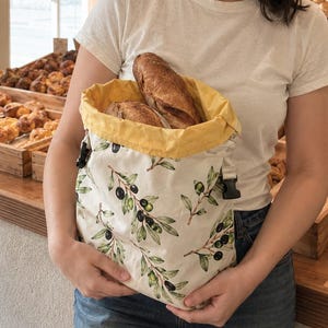 May include: A reusable fabric bread bag featuring a cream-colored exterior with a green olive branch and black olive pattern. The bag has a bright yellow interior lining, a wide opening, and black plastic side-release buckles for closure.