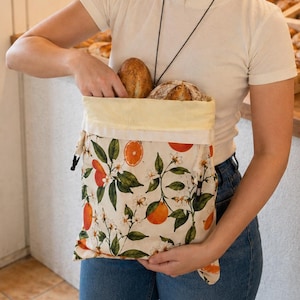 May include: A cream-colored fabric drawstring bag featuring a botanical print of bright orange citrus fruits, green leaves, and white blossoms. The bag is open at the top, revealing two crusty loaves of bread tucked inside the main compartment.