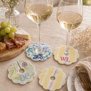 May include: Four scalloped fabric wine glass markers with center slits and circular cutouts. Two feature floral patterns with embroidered initials RS and DC in blue, while two feature yellow stripes with embroidered initials WL in pink.