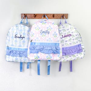 May include: Three personalized backpacks with floral patterns and ruffled accents. The backpacks are white with blue, pink, and purple designs. Each backpack has a name embroidered on it: Evelyn, Emily, and Lauren. The backpacks are hanging on a wooden rack.