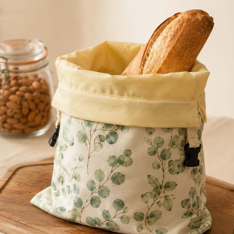 May include: A fabric bread bag featuring a cream-colored base with a green eucalyptus leaf pattern and a solid pale yellow interior lining. The bag has a fold-over top edge and black plastic side-release buckles to secure the opening.