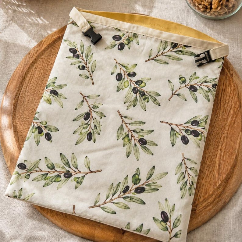 May include: A rectangular fabric pouch featuring a repeating pattern of green olive branches with dark olives on an off-white background. The top edge has a mustard yellow lining and two black plastic side-release buckles for closure.