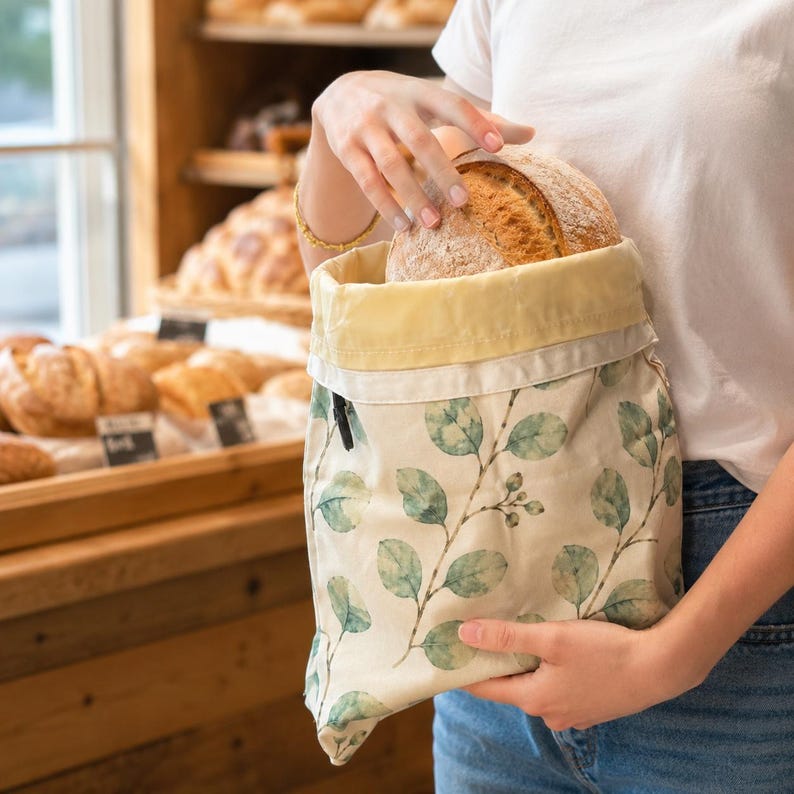 May include: A fabric bread bag featuring a cream-colored base with a green watercolor eucalyptus leaf pattern. The bag has a light yellow interior lining and a drawstring closure, currently holding a round, crusty loaf of artisan bread.