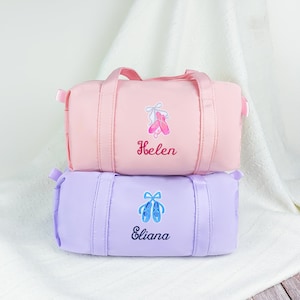 May include: Two small, cylindrical duffel bags, one pink and one lavender, stacked. Each bag has a ballet shoe graphic and a name embroidered below. The pink bag has a ballet shoe graphic and the name "Helen". The lavender bag has a ballet shoe graphic and the name "Eliana".