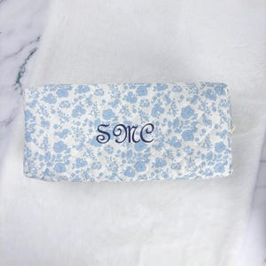 May include: A rectangular, floral-patterned cosmetic bag in white and light blue. The bag features a delicate blue flower print and embroidered initials "SME" in a darker blue font. The bag is resting on a white, fluffy surface.
